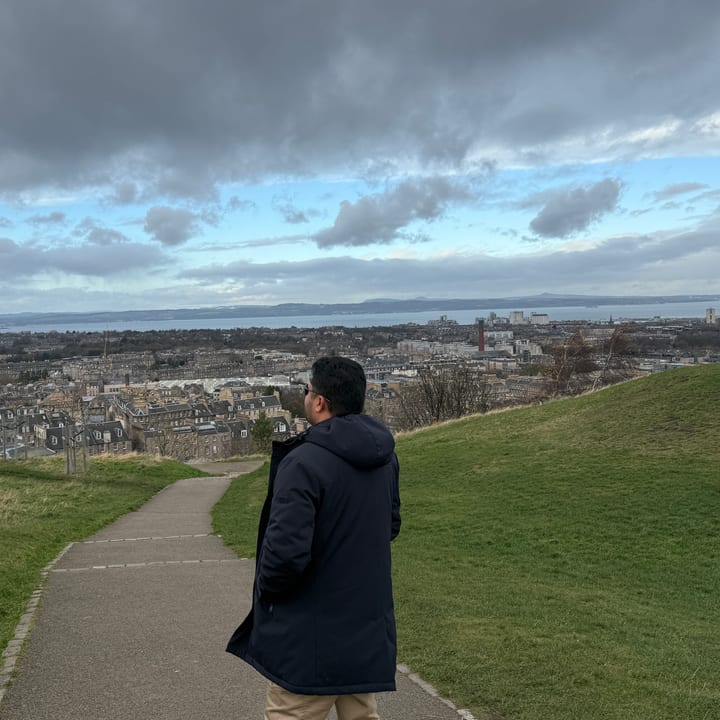 A man in a dark coat stands on a grassy hillside path, looking out over a city of rooftops and a wide estuary beneath a broad cloudy sky.
