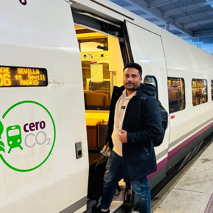 A smiling man with a backpack stands in the open door of a white high-speed train at a station platform; the destination display reads Sevilla and a green 'cero CO2' logo is on the side.