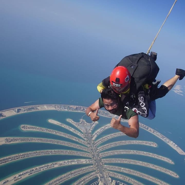 Two people in tandem freefall above the sea, the one in front wearing a red helmet, with a palm-shaped island below them and a hazy blue horizon.
