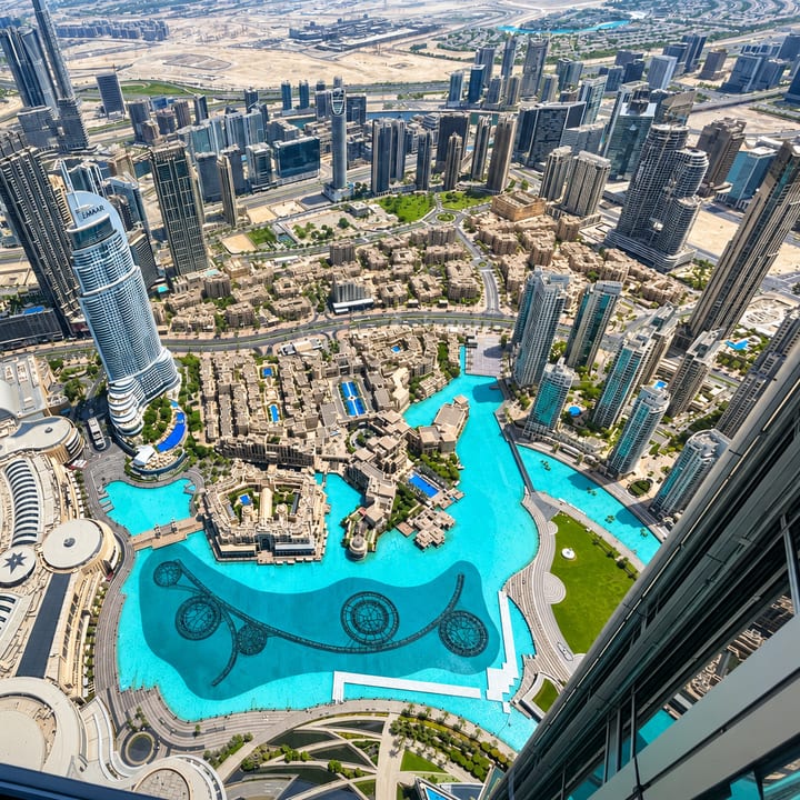 Looking straight down from a very tall tower onto Downtown Dubai: glass skyscrapers, low sand-coloured buildings and a bright turquoise lake with a dark, patterned fountain basin.