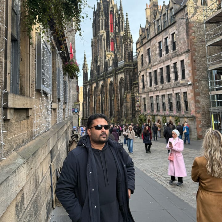 A man in sunglasses and a dark jacket stands on a paved old-town street with a tall Gothic church spire and weathered stone buildings behind him and pedestrians in the distance.