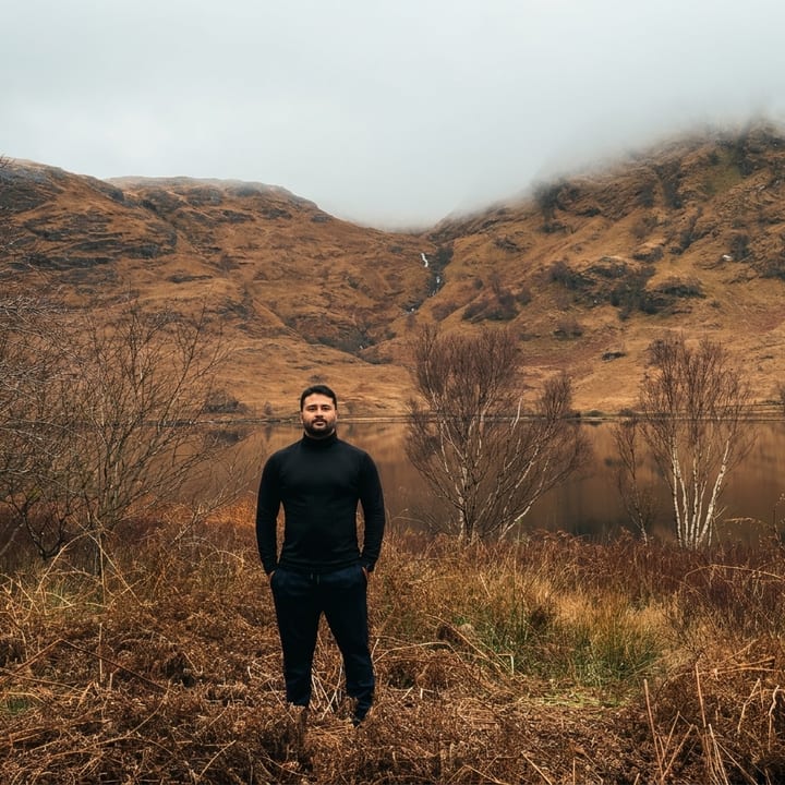 A man in black stands among golden-brown bracken beside a still loch, with bare birch trees and misty, rust-coloured hills rising behind him under a pale grey sky.