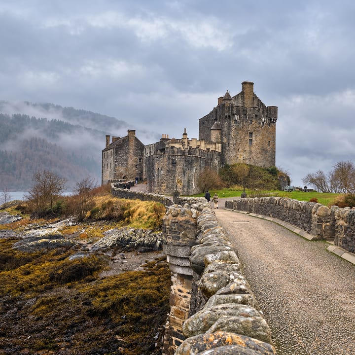 A stone bridge with low walls leads across a rocky shore to Eilean Donan Castle, a grey stone castle with several towers, set against misty wooded hills and a cloudy sky.
