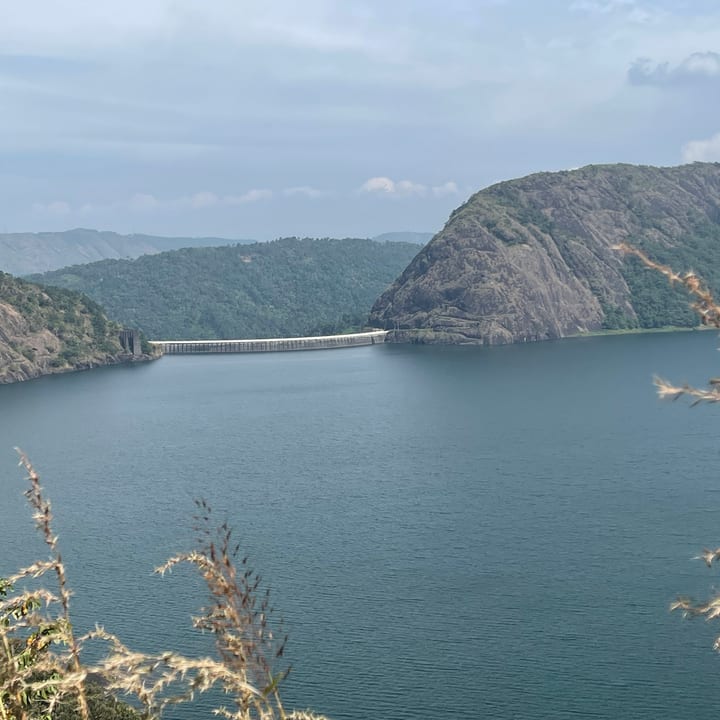 A wide blue-green reservoir held between two rocky, forested hills by a slender concrete dam, with layered ridges beyond and dry grasses in the foreground.