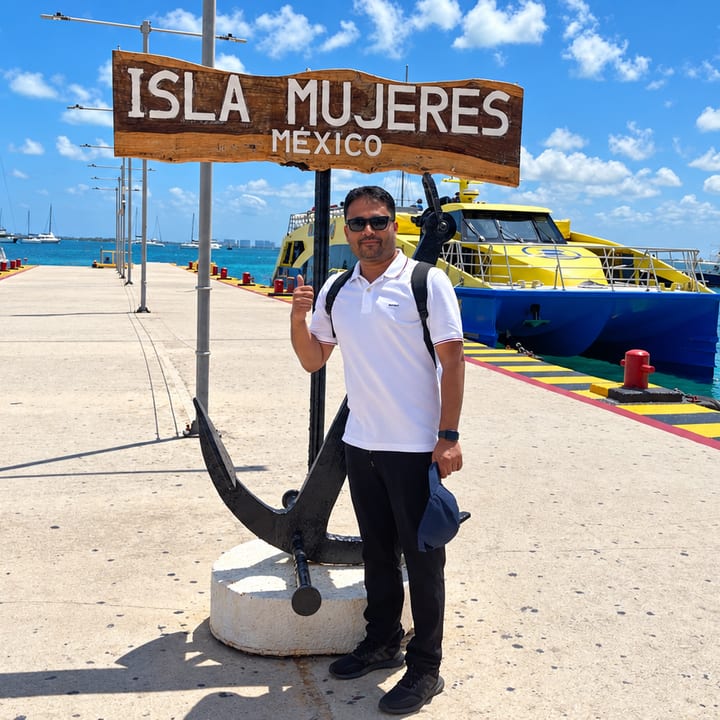 A smiling man in a white shirt, a cap in his hand, stands beside a dark anchor on a long concrete pier under a wooden 'Isla Mujeres México' sign, with a yellow and blue ferry and turquoise water beside him.