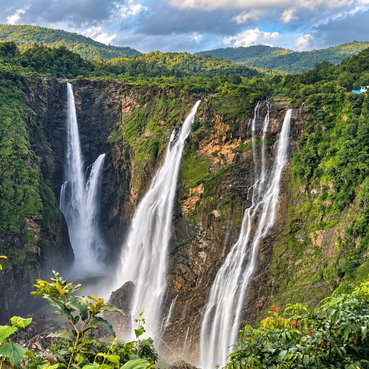 Several tall waterfalls plunge down a steep gorge of rock and dense green forest, with lush leaves in the foreground and scattered clouds above.