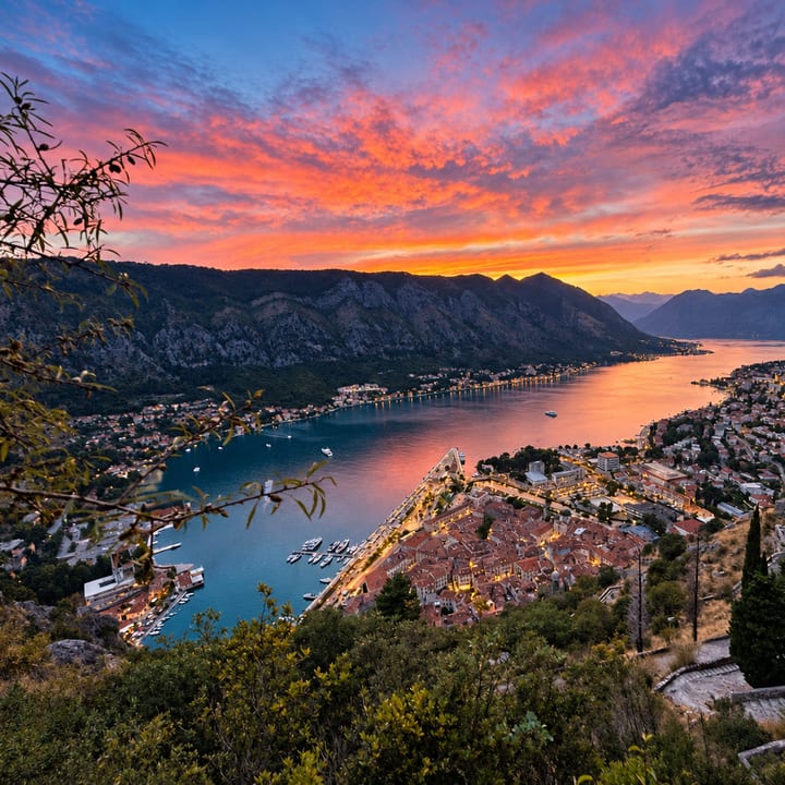 A fiery orange and pink sunset sky over a bay, with a walled old town of red roofs below, the water glowing and layered mountains stretching into the distance.