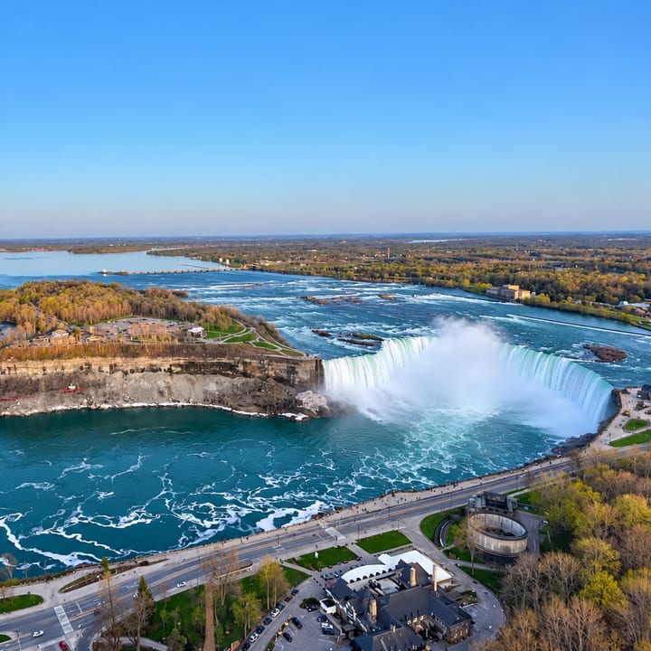Aerial view of Niagara Falls: two broad waterfalls drop into a turquoise river gorge beside a green riverside road, with a town and wide river beyond under a clear blue sky.
