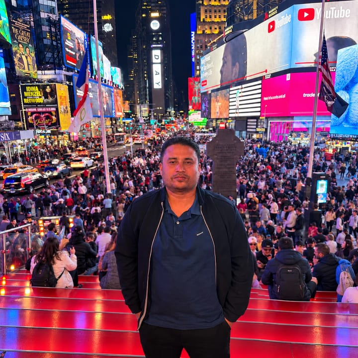 A man in a dark jacket and blue polo shirt stands on glowing red steps at night, with a dense crowd and the bright billboards of Times Square behind him.