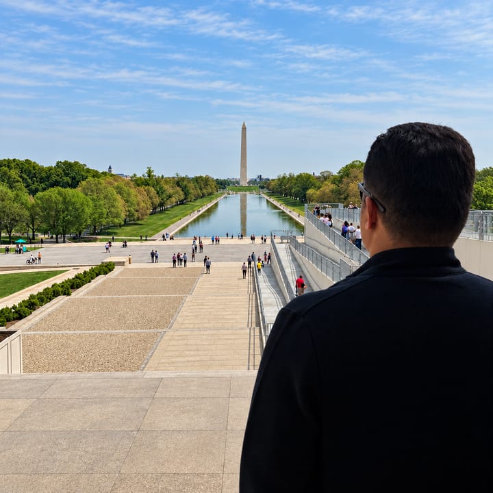 A man seen from behind looks along a long reflecting pool towards the Washington Monument, with rows of trees on either side and a blue sky with thin clouds.
