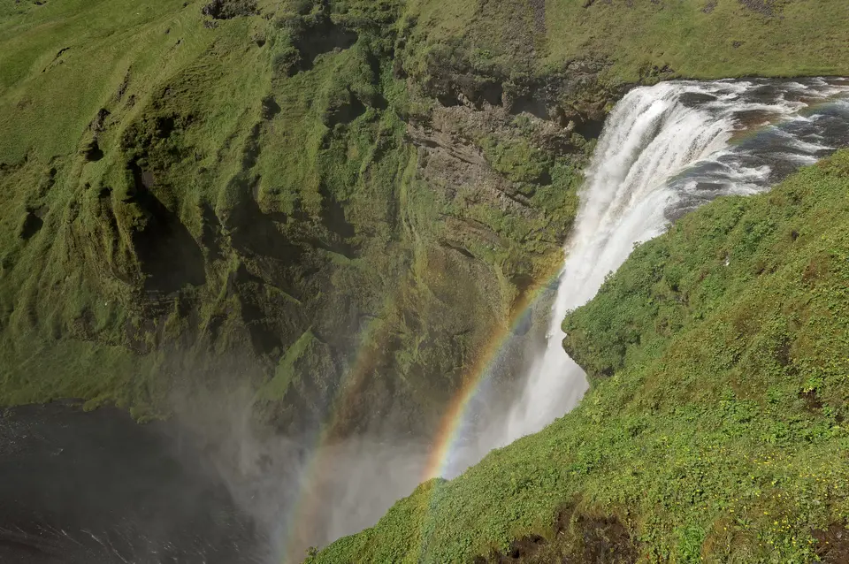 The top of a waterfall with a rainbow in mist and green slopes beside it.