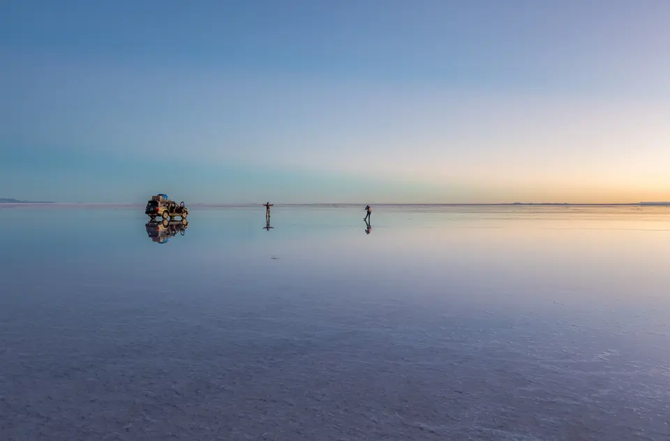 Sunrise over the flooded Salar de Uyuni, with a 4x4 vehicle and two people reflected in the still water.