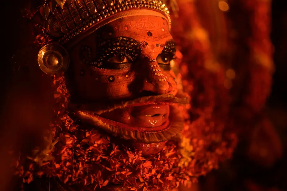 Close-up of the red face paint, beaded eye decoration and flower garlands of a Kandanar Kelan Theyyam performer, lit by firelight.