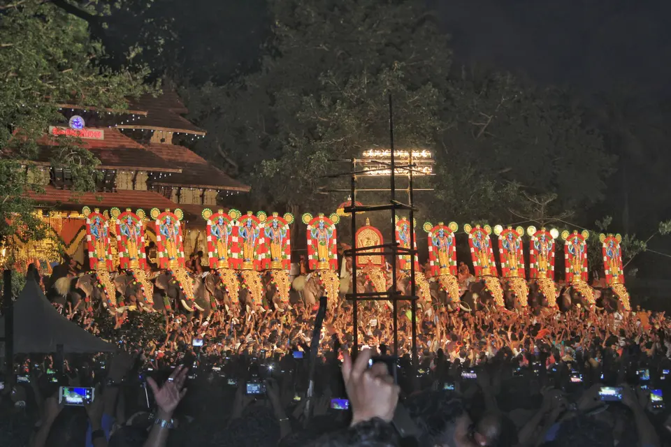 Caparisoned elephants with ornamental nettipattam lined up at night during the Kudamattam at Thrissur Pooram, with a dense crowd in front.