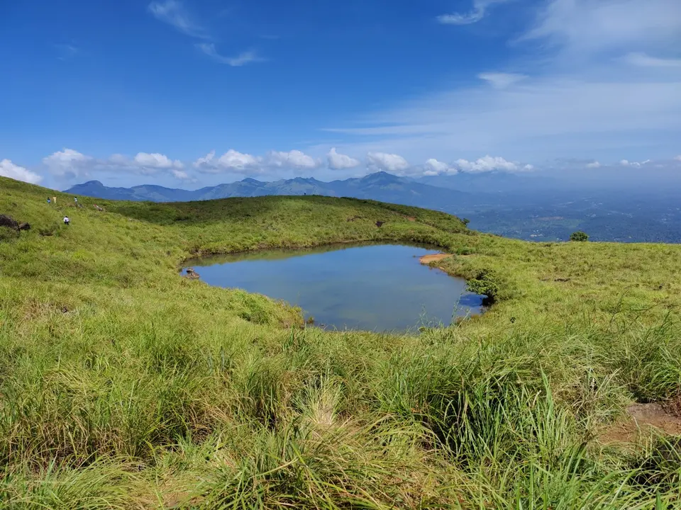 A small lake called Heart Lake on a grassy hill near Chembra Peak in Wayanad under a blue sky.