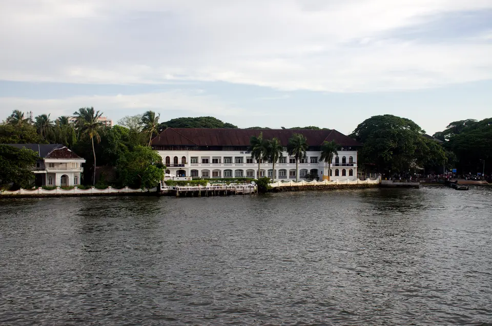 The white colonial-style Brunton Boatyard hotel building seen from the water with palms and a cloudy sky in Fort Kochi.