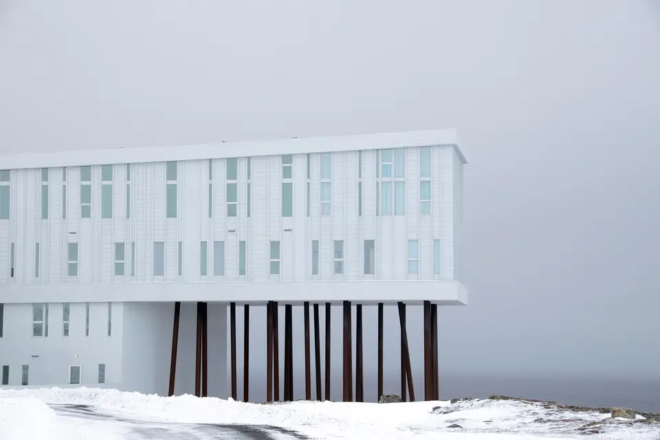 A white multi-storey inn raised on dark wooden stilts above rocks and snow in grey mist on Fogo Island.