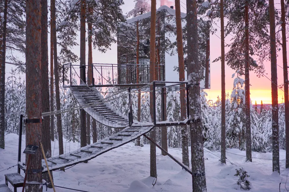 A staircase leading up to a mirrored cube suspended among snowy pine trees at sunset at Treehotel in Harads, Sweden.