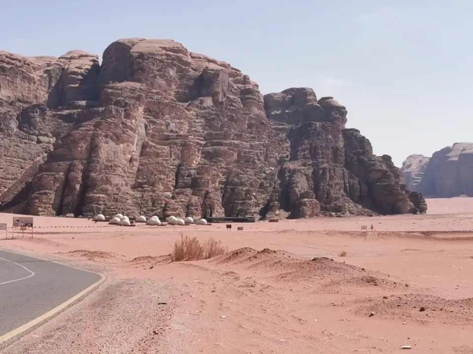 Pale tents beside a road and a sandy plain at the foot of red sandstone cliffs in Wadi Rum.