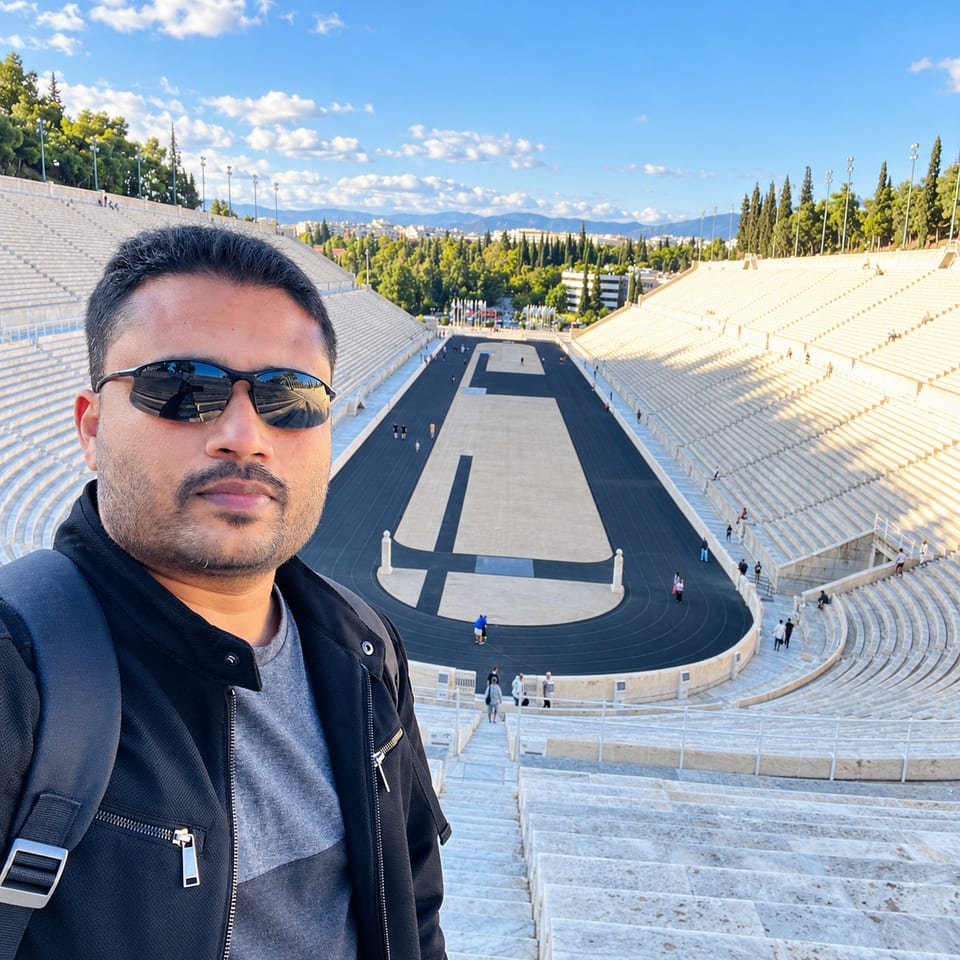 A selfie of the traveller in sunglasses high in the stands, with the stadium's track, the trees and the city behind.