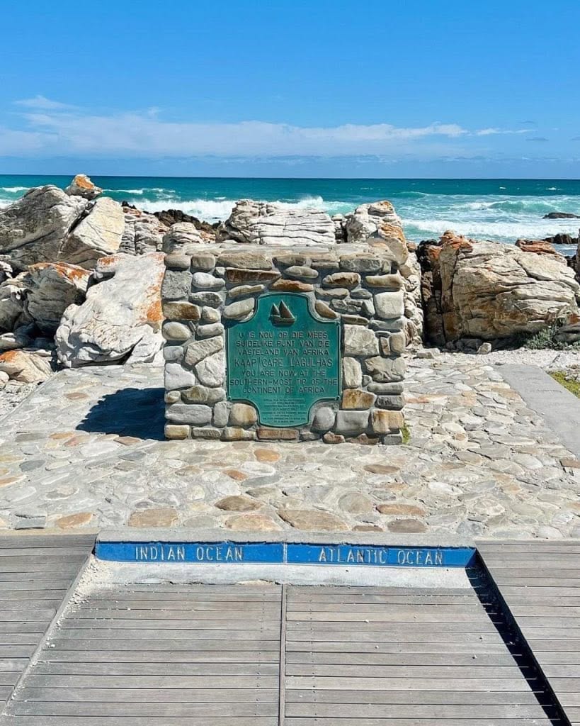 The stone cairn with a green plaque marking the southernmost tip of Africa; "Indian Ocean" and "Atlantic Ocean" are painted on the kerb in front, with waves breaking on the rocks behind.