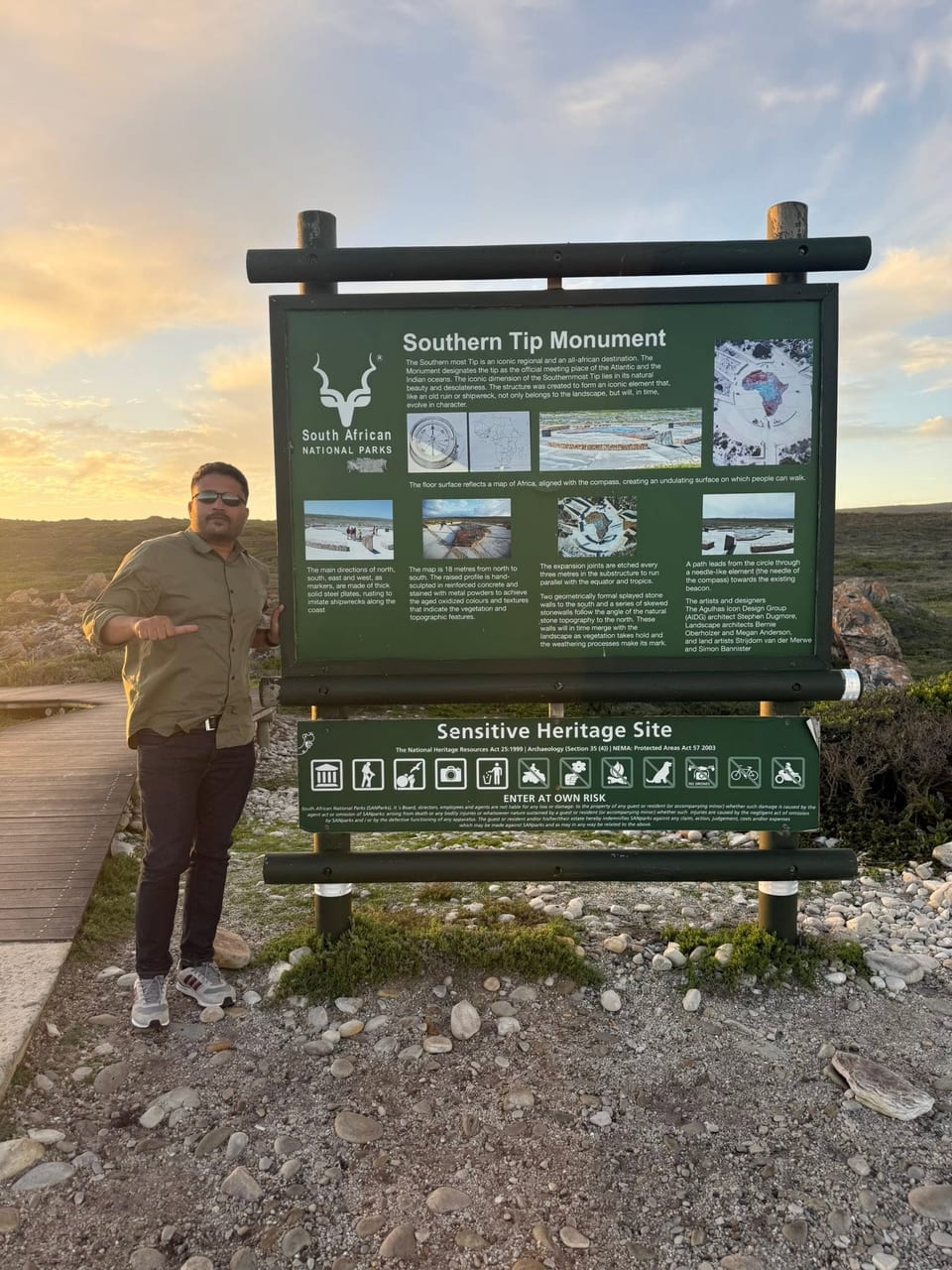 The traveller stands beside the South African National Parks "Southern Tip Monument" information board at sunset.
