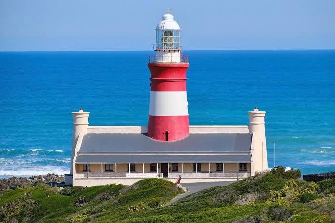 The red and white Cape Agulhas lighthouse tower rising from a cream-coloured building, with the blue sea behind.
