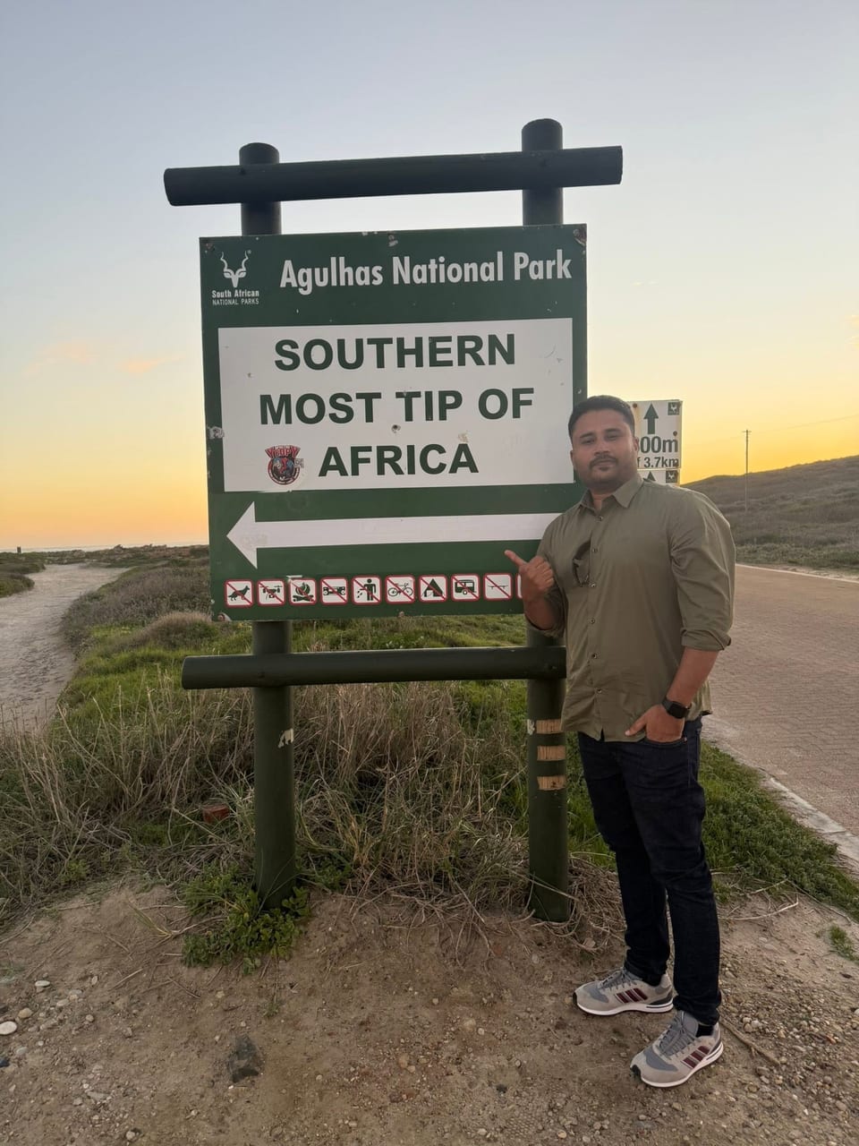 A traveller in an olive shirt points to the Agulhas National Park sign reading "Southern Most Tip of Africa" at sunset.