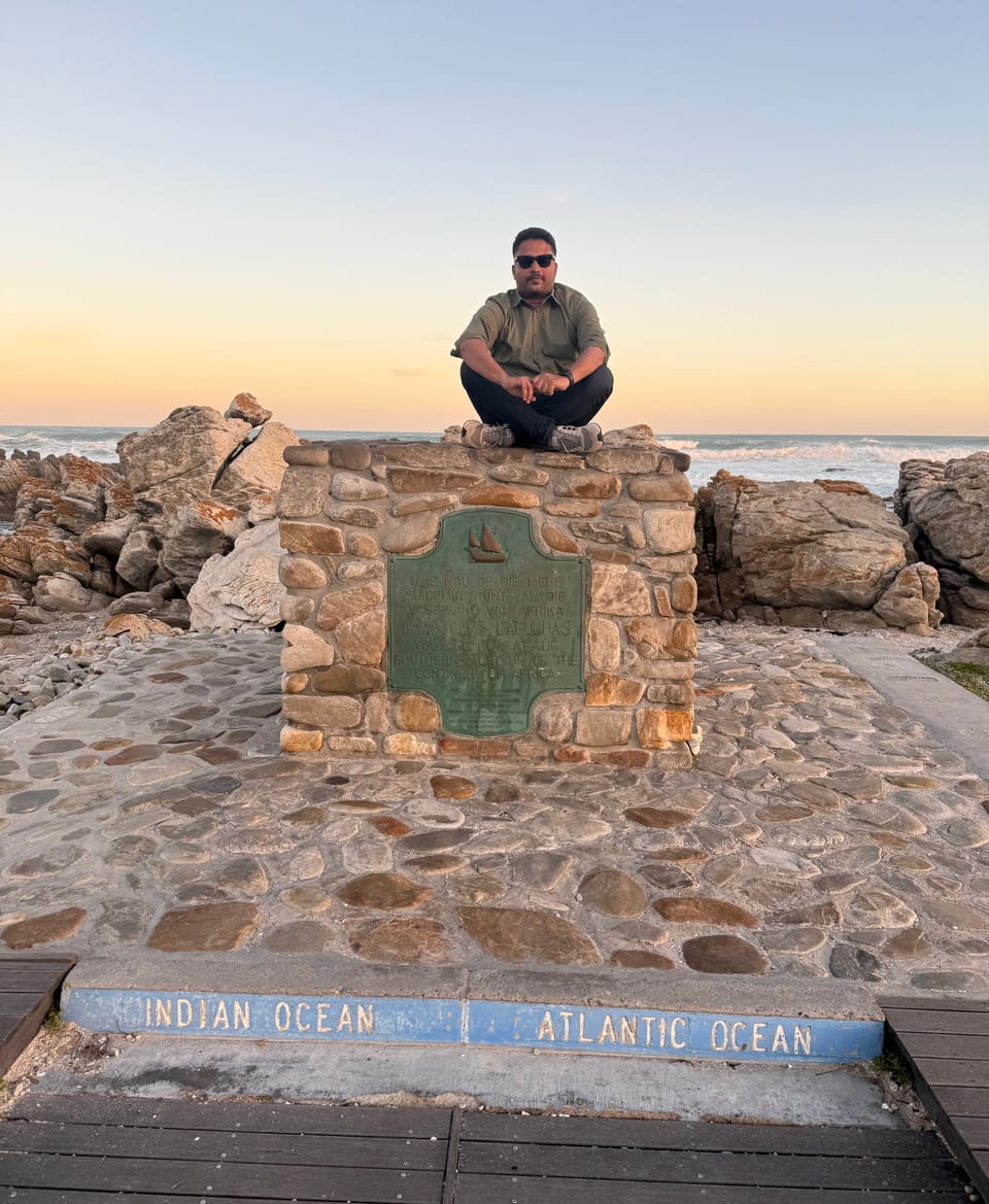 At dusk, the traveller sits cross-legged on top of the stone cairn at the southern tip of Africa, above the painted "Indian Ocean" and "Atlantic Ocean" marker, with the sea behind.
