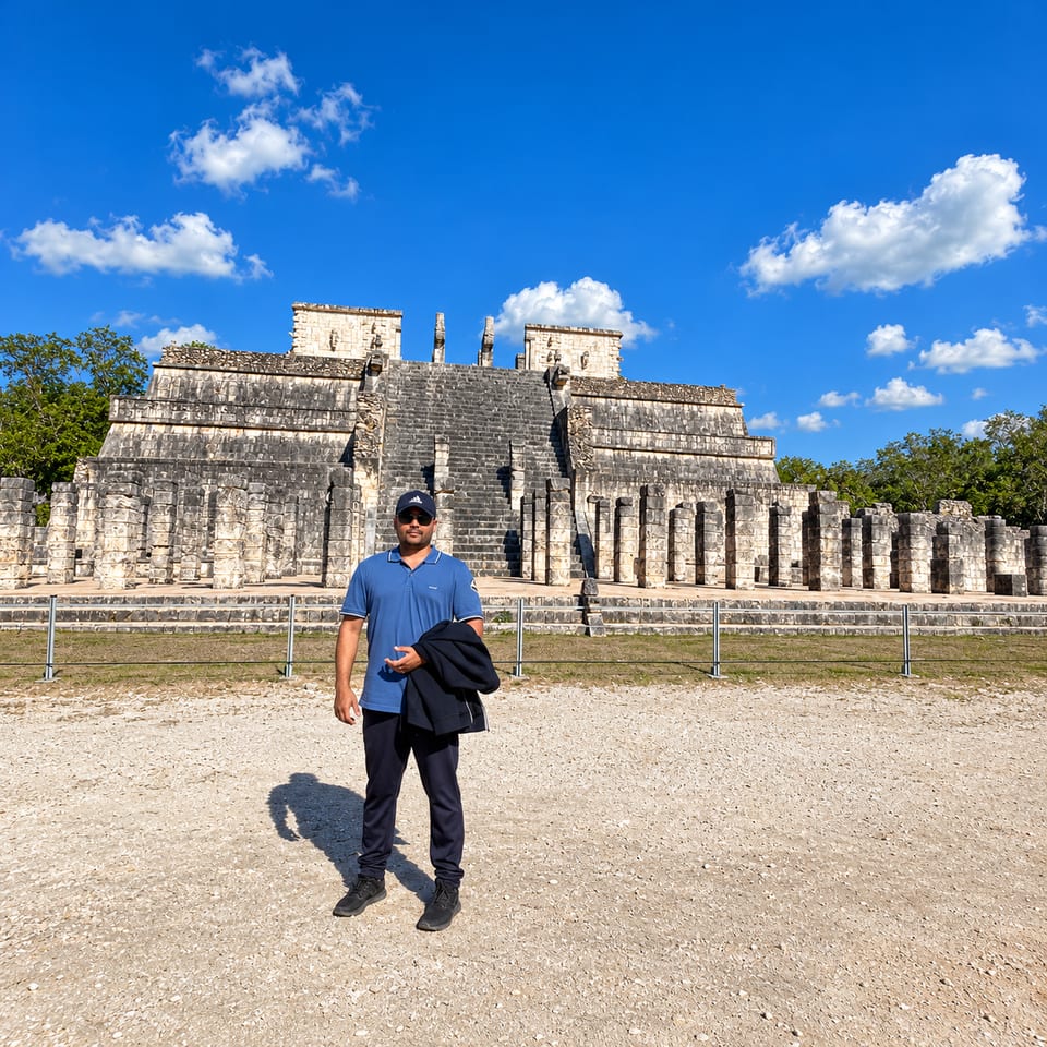 The traveller in a cap stands on a gravel path in front of the Temple of the Warriors, with rows of square stone columns along its base.