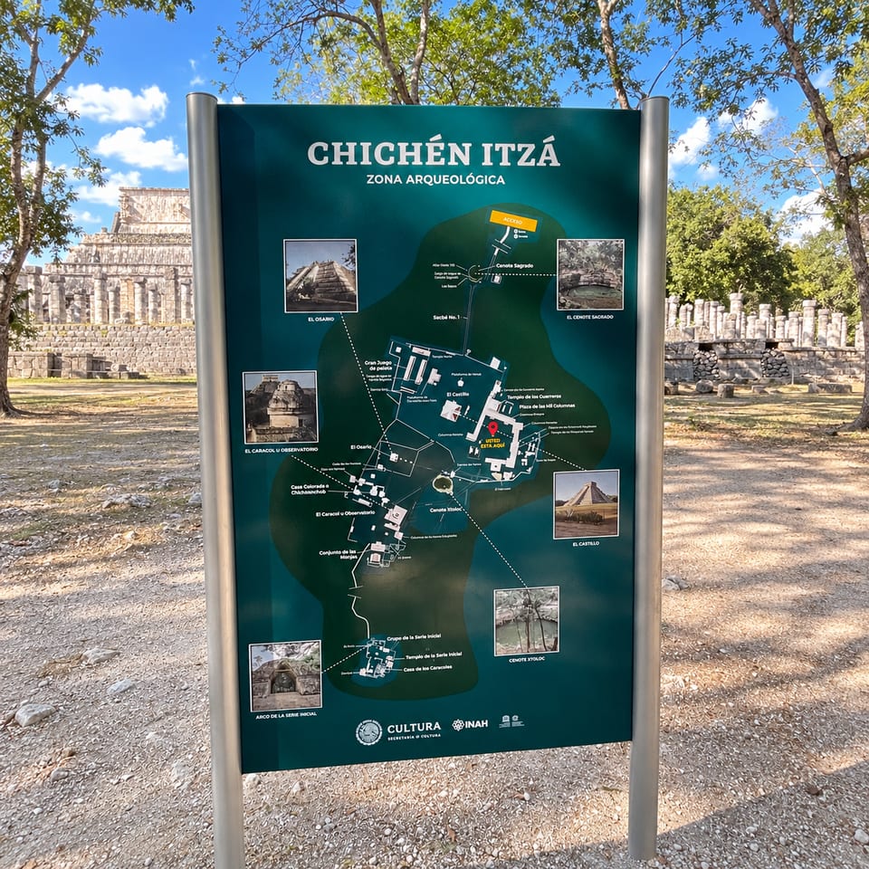 A green information board titled "Chichén Itzá, Zona Arqueológica" with a map of the site and photographs of its main buildings, standing among trees.