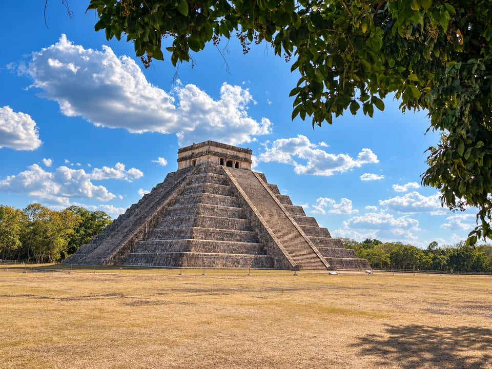 El Castillo seen across a dry grass field, framed by the leaves of an overhanging tree.