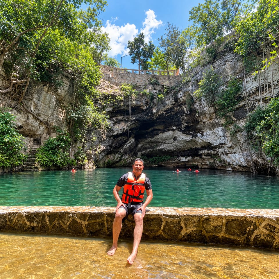 A man in an orange life jacket sits on a stone ledge in shallow water at the edge of a cenote, a deep natural pool ringed by rock walls and trees, with swimmers behind him.